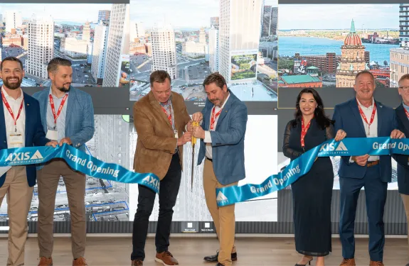 Five people holding a grand opening ribbon while two in the middle use giant scissors to cut it.