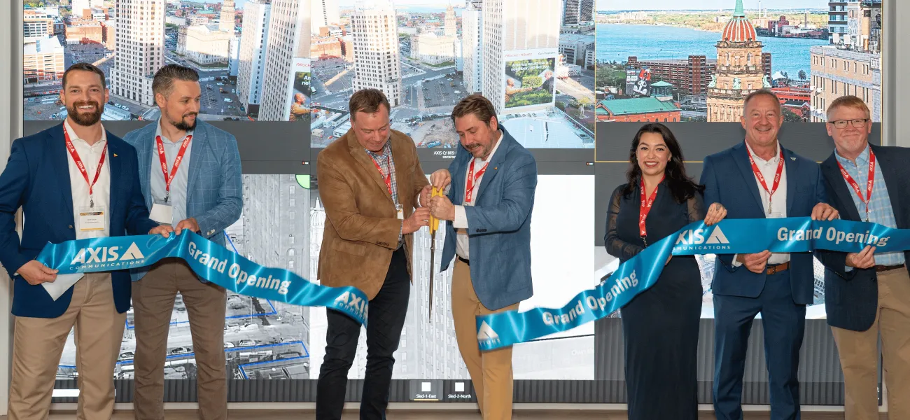 Five people holding a grand opening ribbon while two in the middle use giant scissors to cut it.