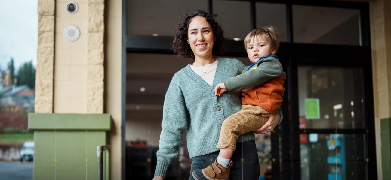 A woman stands outside of a retail store holding her son with an Axis camera in the background